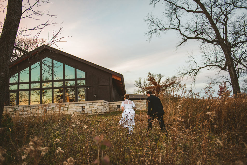 a bride and groom walking back to their reception in the fall at sunset with the pink sky in the background
