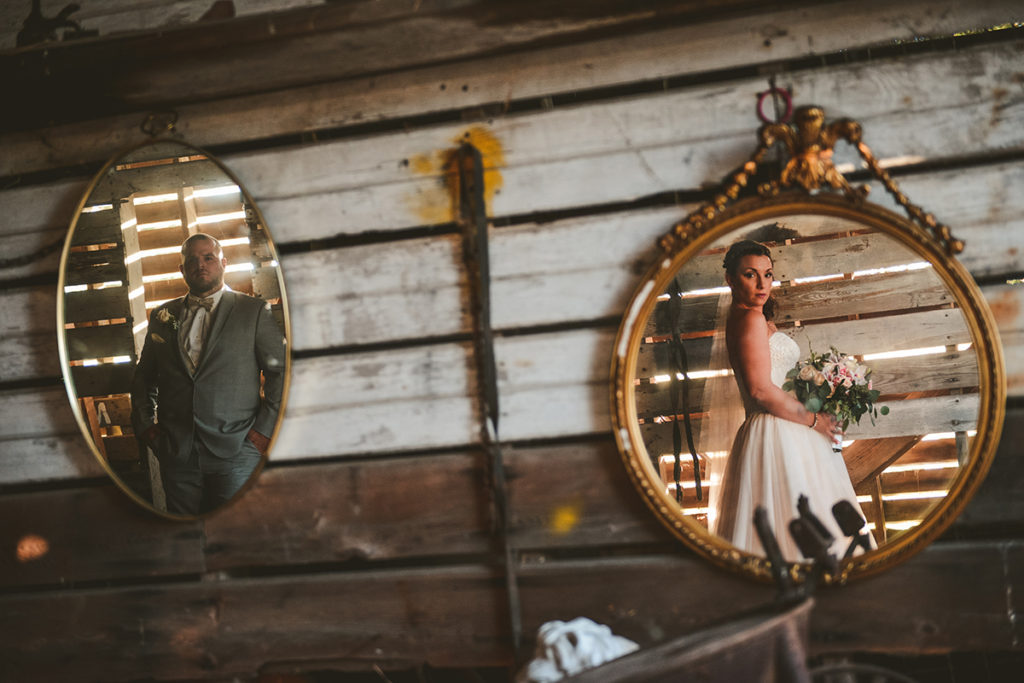 a bride and groom standing in an old barn as they look into two vintage mirrors