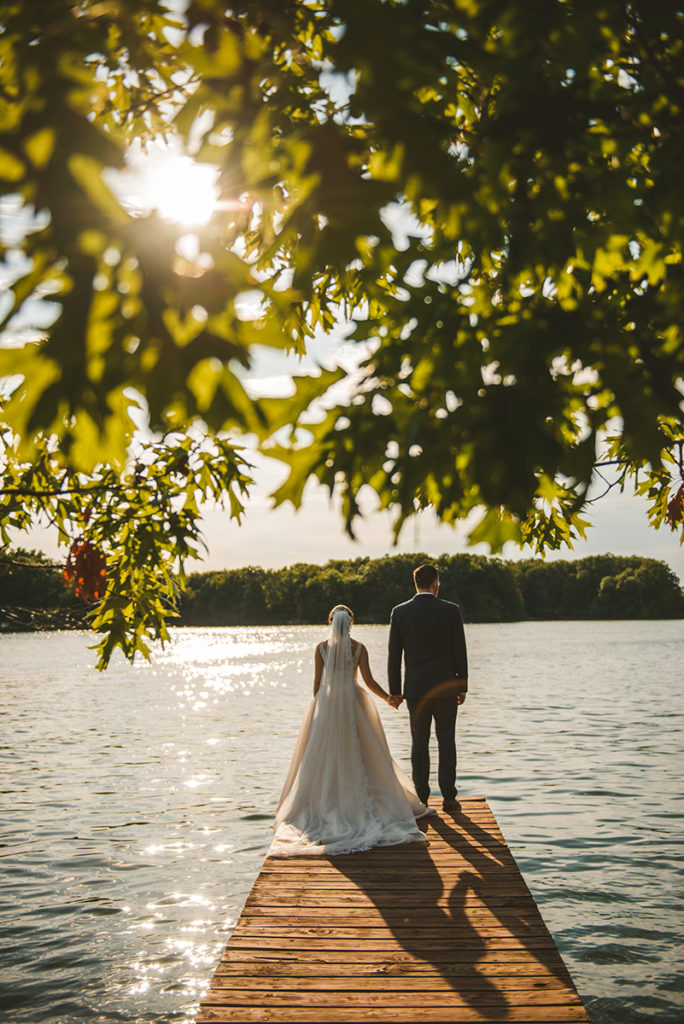 a recently married couple standing on a old dock as the sun sets over the lake