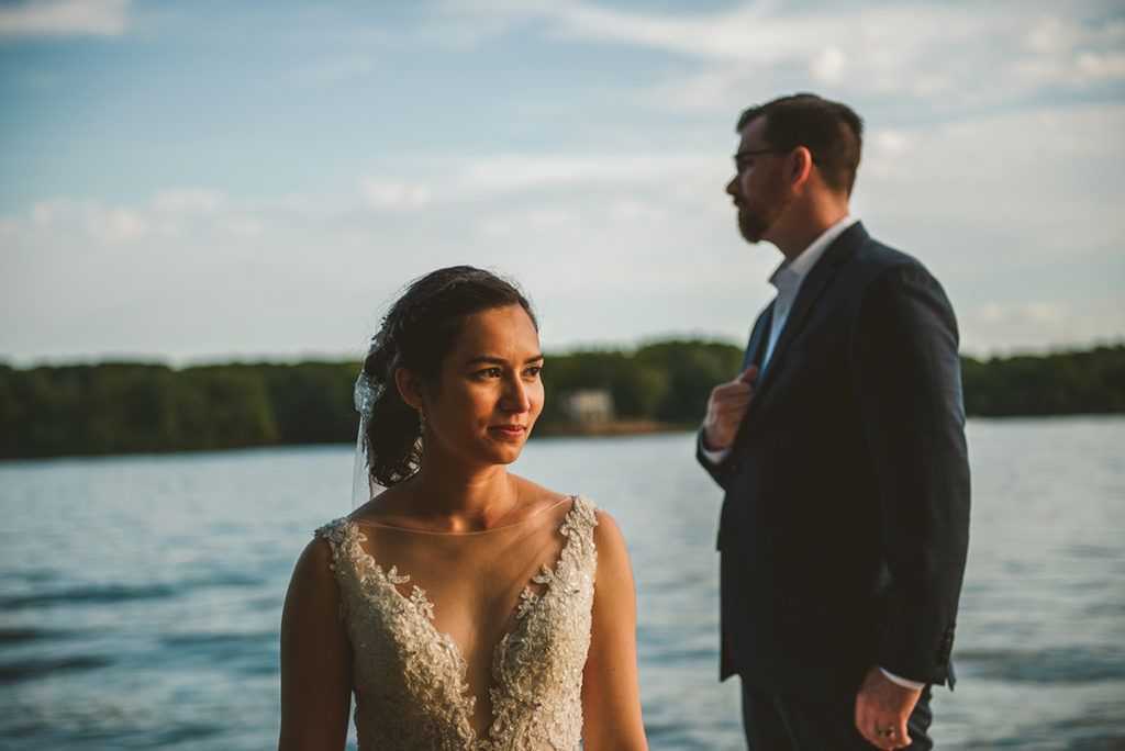 a beautiful bride in a gorgeous vintage dress stands in the setting sun with groom standing over her in the background