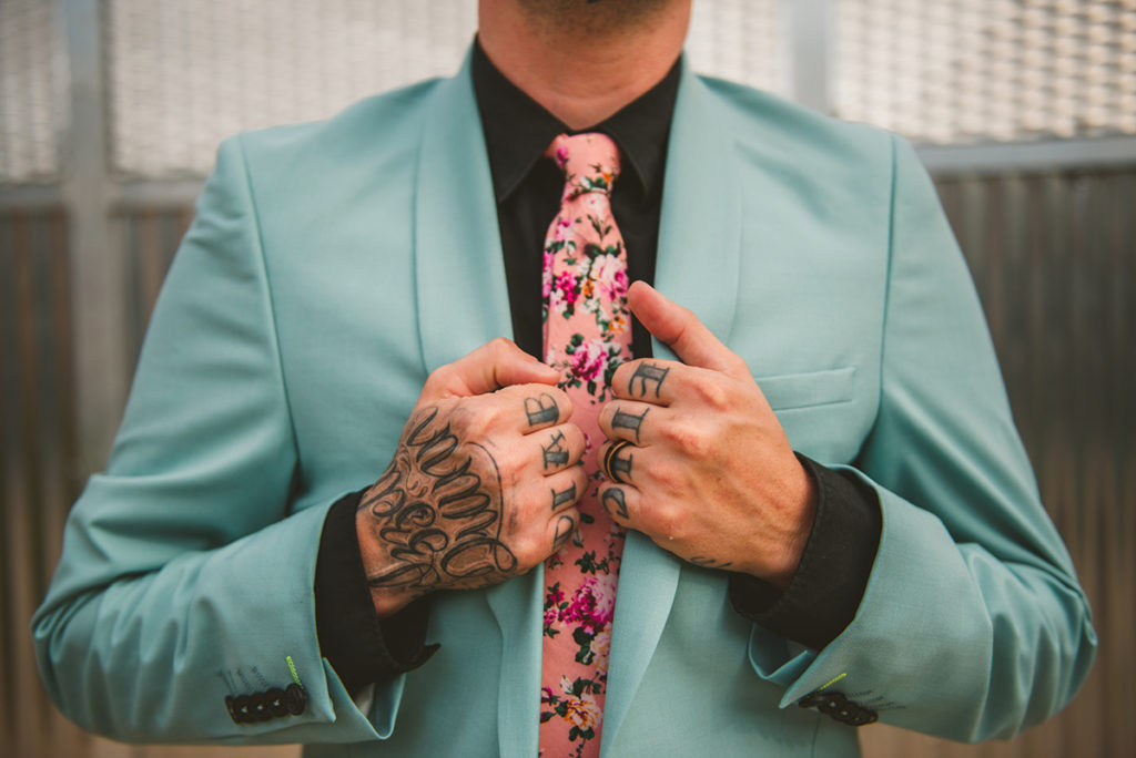 a tattooed groom standing in a baby blue suit as he fixes his jacket and pink tie