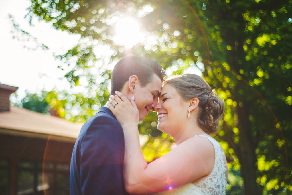 a newly married bride and groom laughing as they stand together as sunlight cascades through the trees in summer