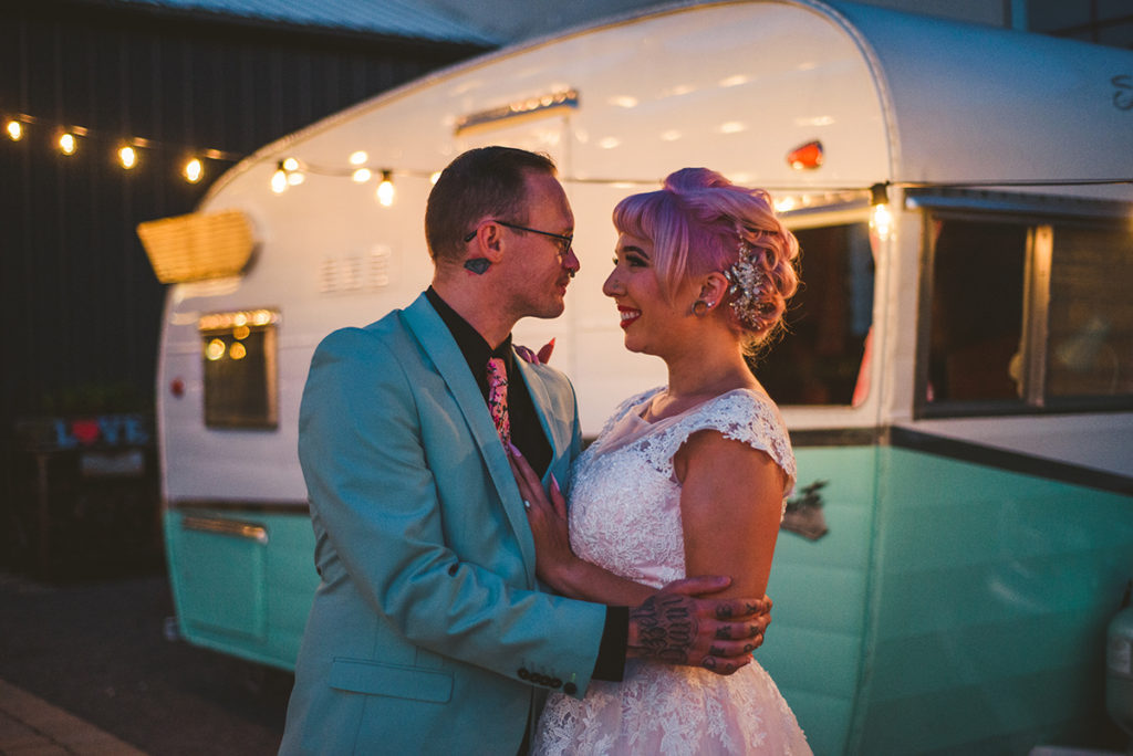 a tattooed wedding couple standing in front of a old camper in the evening of their wedding