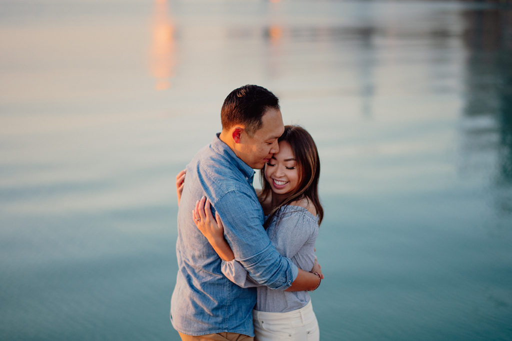 a husband and wife standing close together as they hug in front of pastel blue water at sunrise