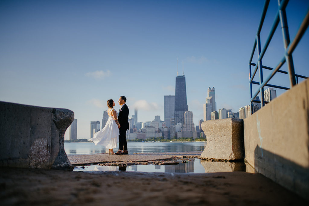 a woman in a white flowing dress standing with her husband in front of the Chicago skyline at North Avenue Beach at sunrise