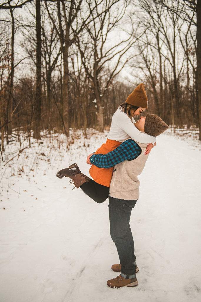 a young engaged couple playing the snow as they laugh in the woods
