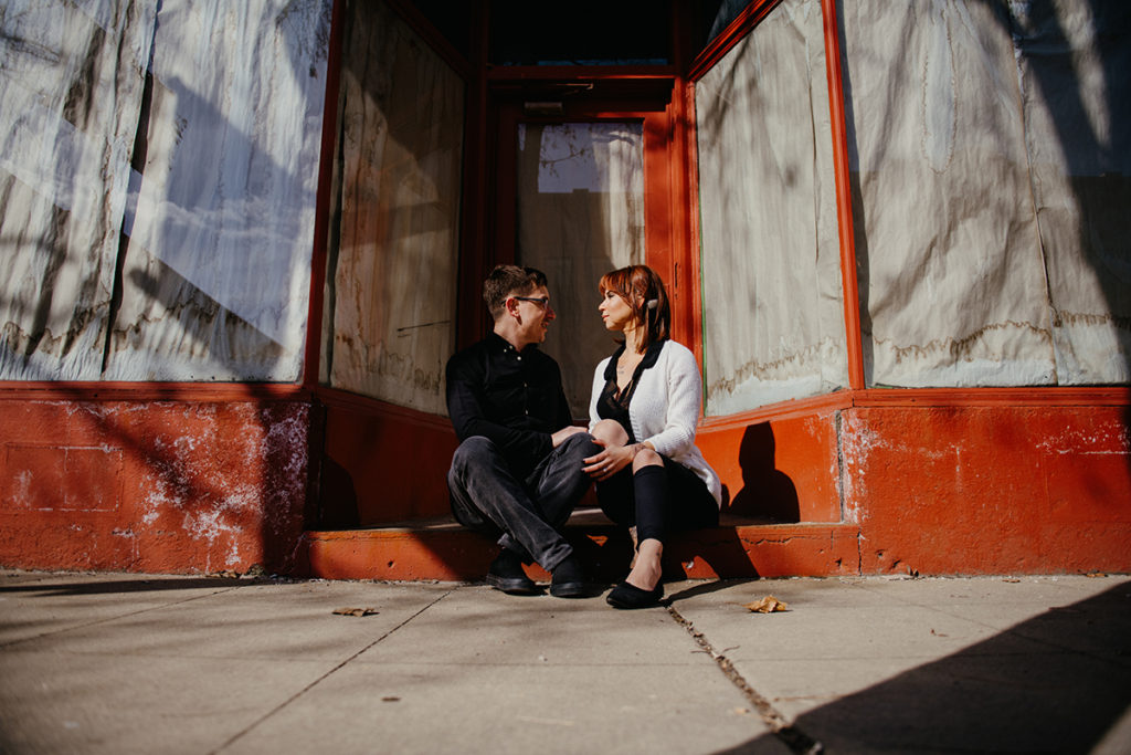 an engaged couple getting close as they smile at each other as they sit in front of an old building