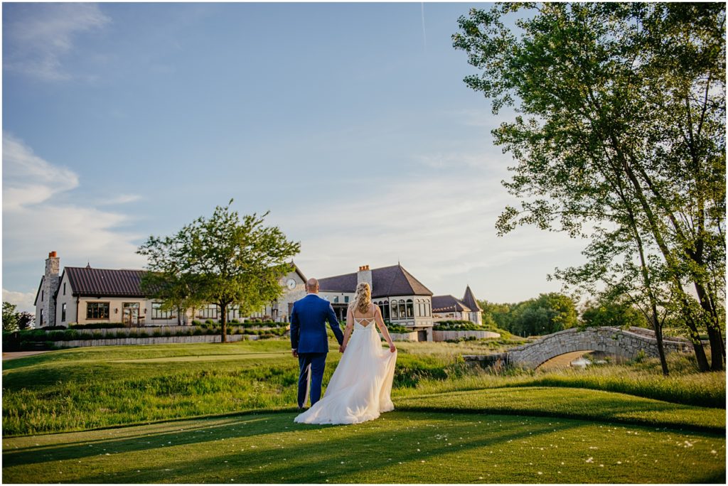 a bride and groom holding hands as they look at Mistwood Golf Club in the distance during golden hour on a warm summer night