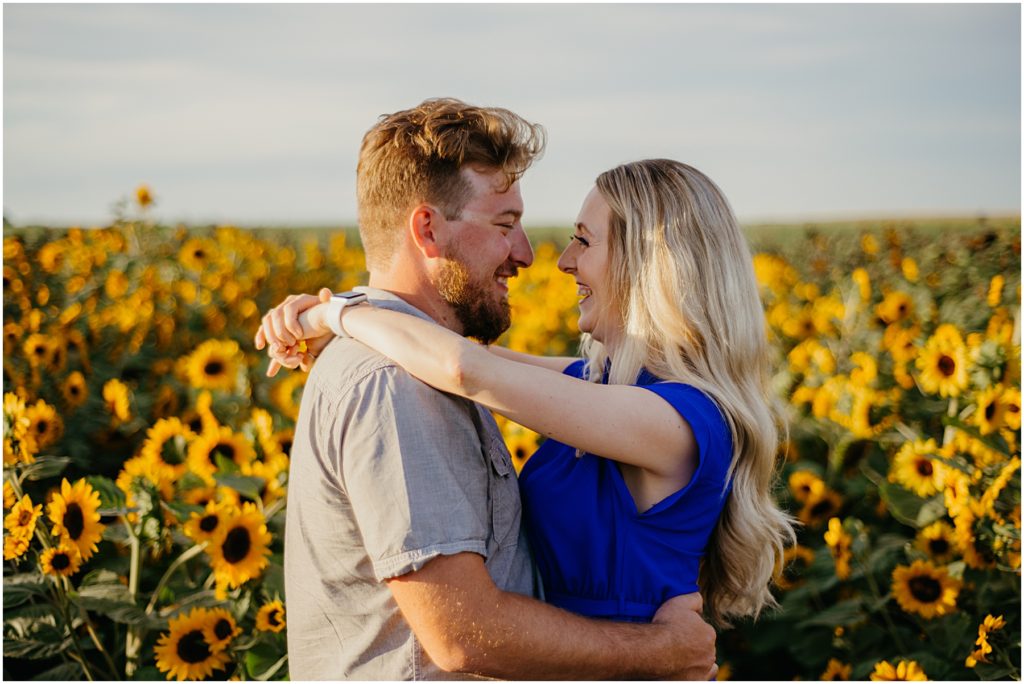 a husband and wife laughing as they go in for a kiss while standing in a golden field of sunflowers