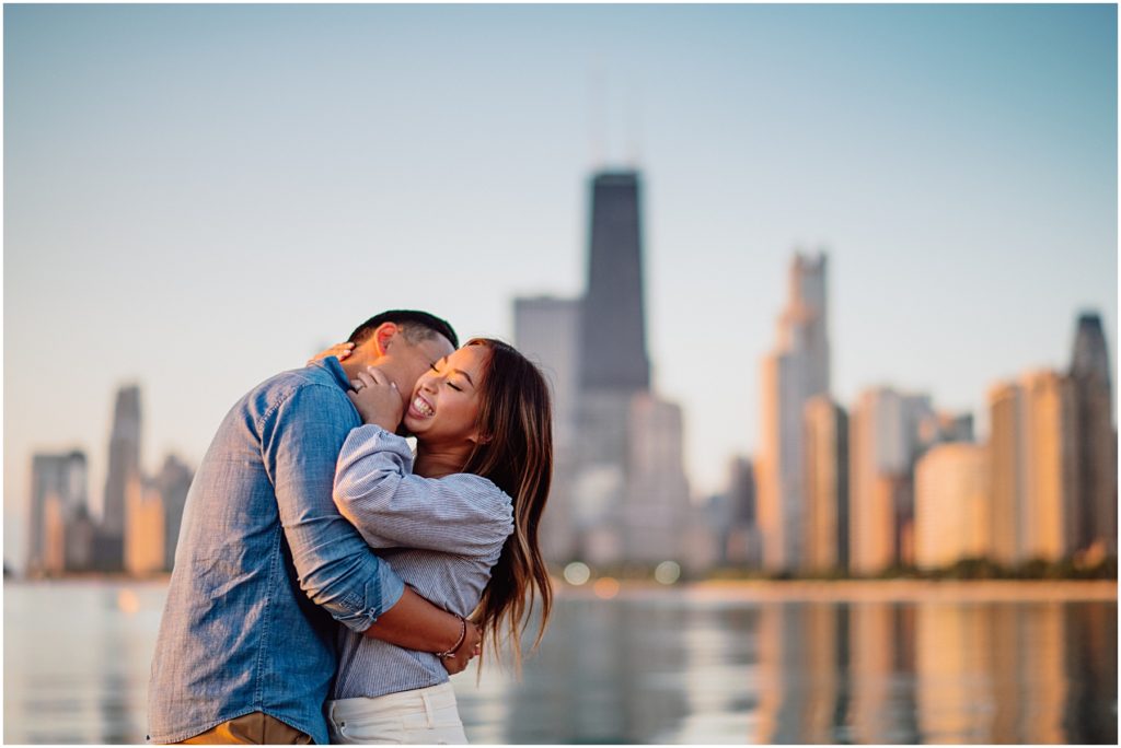 a picking a photographer out of the many Chicago engagement photographers out there a man kisses his finaces neck in front of the golden Chicago skyline at their North Avenue Beach sunrise engagement session