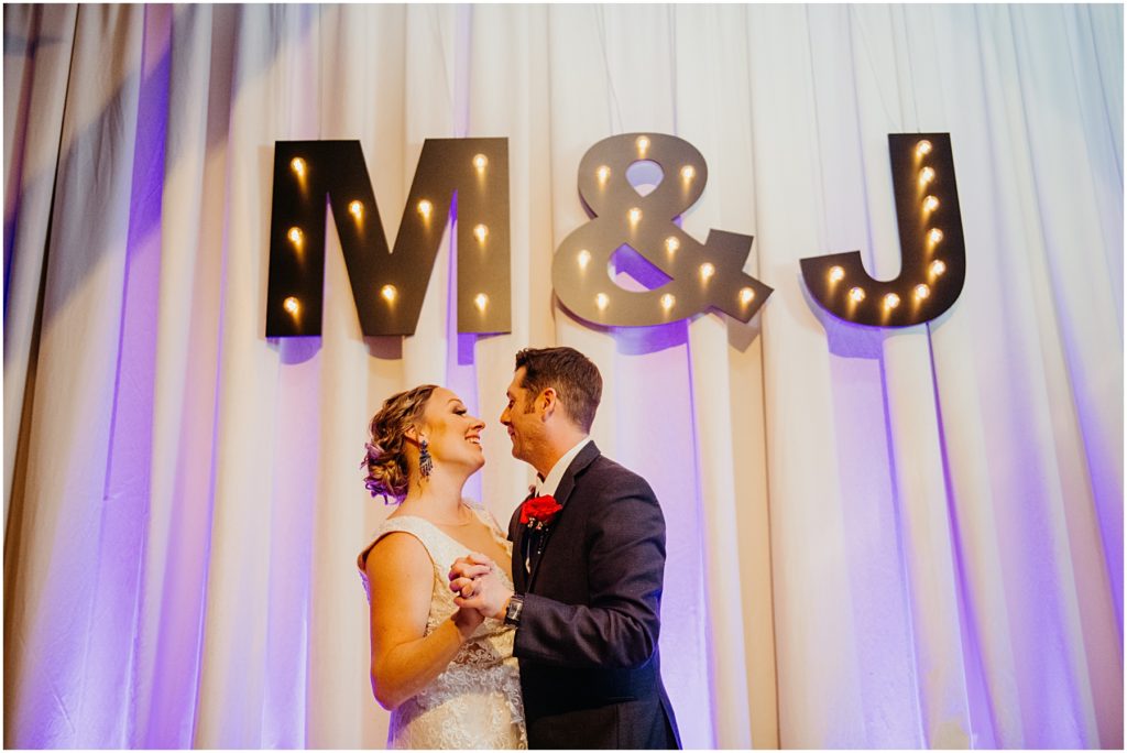 the husband and wife laughing as they dance under a M & J at their warehouse wedding