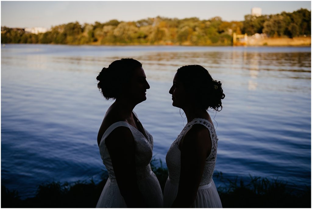 two brides standing silhouetted against a river at sunset 